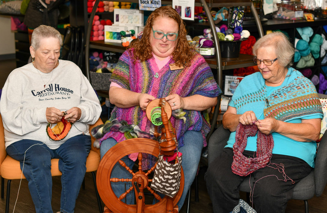 3 women sitting in a row. the two on the outside are knitting, the one in the center is spinning yarn on a spinning wheel