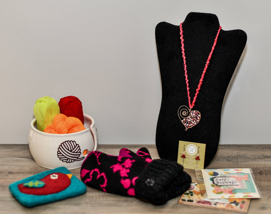 A collection of artisan creations including a yarn bowl, a heart-shaped beaded necklace, pink and black mittens, a felted pouch and greeting cards